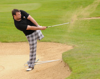 a male golfer swings his club pivoting on one leg to drive the ball out of a bunker