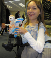 A photograph of Esther Apleyard holding both the Mascots for the Olympic and Paralympic Games.  They are soft toys, about 15cms heigh mainly in blue and white and orange and white.  They have helmet type shapes on their heads and one central eye which represents a camera